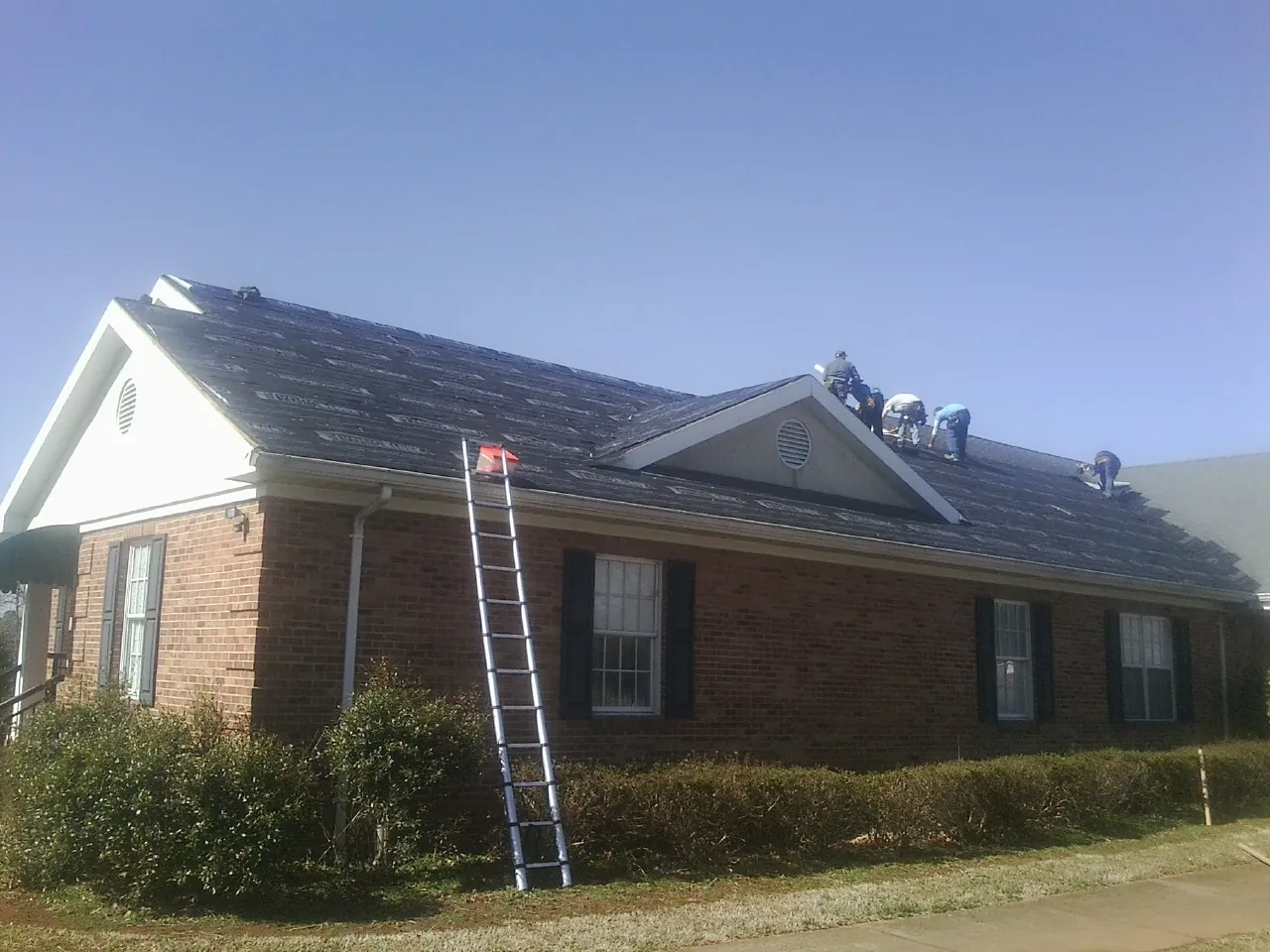 Crew tearing shingles off a brick ranch roof with a ladder set — Emergency Roof Repair work by our crew in Roaring Spring